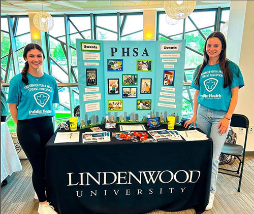 Two St. Louis public health degree students wearing teal shirts standing at a table presentation of their degree program
