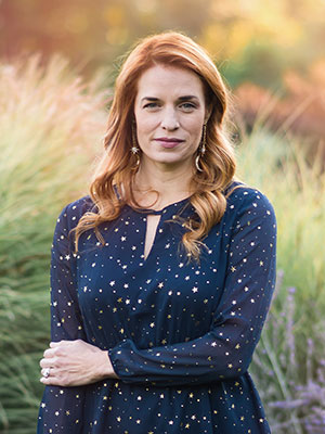 White woman with red hair standing in a field wearing a deep teal blouse with gold stars
