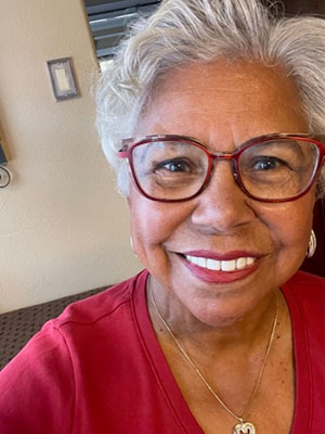 Older woman with brown skin and grey hair smiling wearing a red shirt and red glasses
