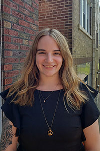 woman with blonde hair wearing a black shirt with a tattoo sleeve standing against a brick building