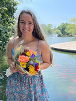 White woman with long dark blonde haor holding flowers in front of river