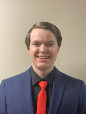 White man with light brown hair wearing a black dress shirt and navy blue suit jacket with a red tie