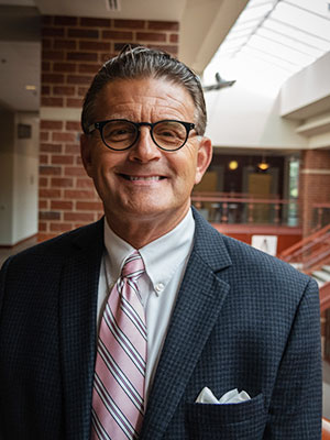 Harry H. (Chip) Peterson Older man with dark slick backed hair wearing glasses a dark grey suit and pink stripped tie standing in Spellmann Center