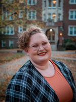 Girl with short curly hair wearing glasses and a plaid shirt smiling standing infront of campus building