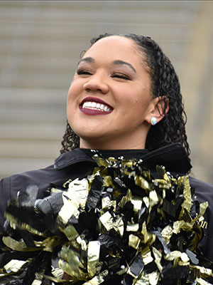 Smiling young lady with brown skin wearing a black long sleeve jacket holding cheer pom poms. She has short black curly hair.