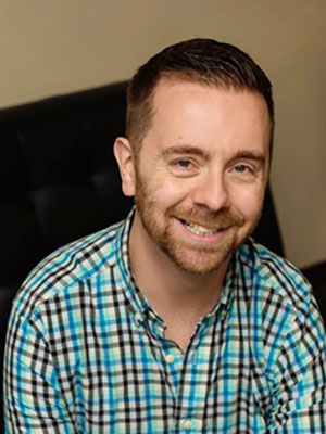 Man with brown hair and beard wearing a light blue and dark brown plaid shirt