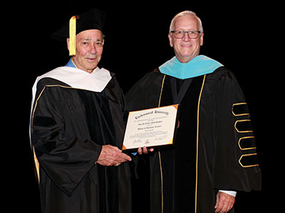 Jerry Scheidegger wearing black doctoral robes with white hood, being handed his honorary doctorate by President Porter wearing black doctoral robes with blue hood.