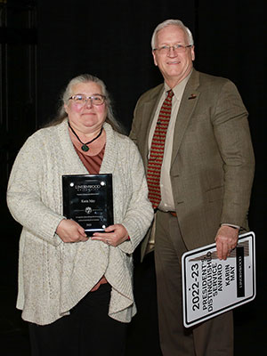 Woman with grey straight hair behind her shoulders wearing a beige sweater and brown shirt holding an award. Man with short grey hair wearing a brown suit and red tie holding the parking spot sign for the award recipient.
