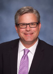 White man wearing glasses, a black suit, and purple tie