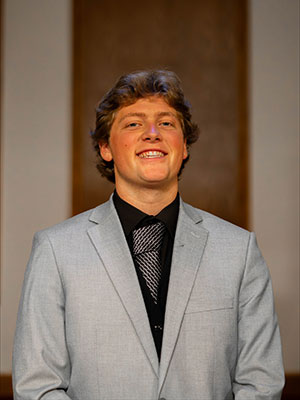White man with light brown hair wearing a black dress shirt and grey suit jacket
