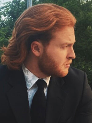 White man with red hair and a red beard wearing a black suit