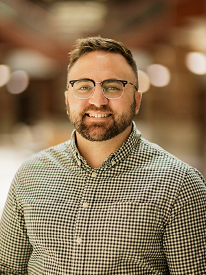 White man, brown hair brown beard, light colored dress shirt standing outside infront of trees