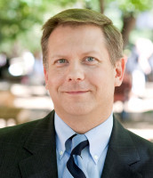 White man with blonde hair and blue eyes wearing a dark suite with a light blue shirt standing outside